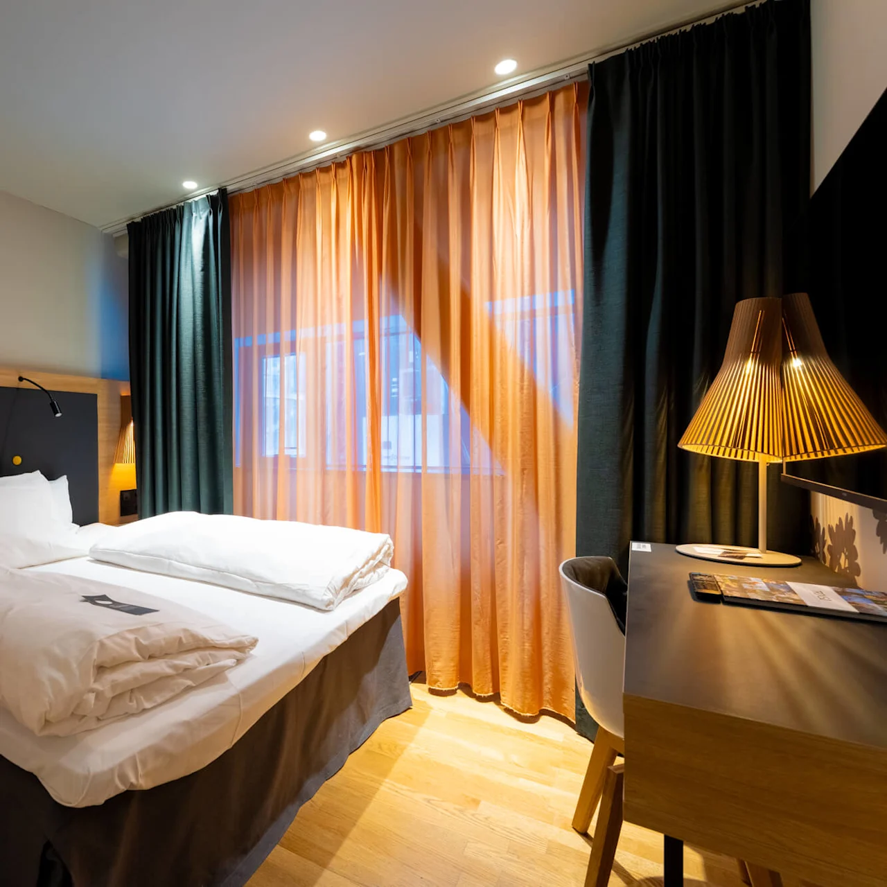 En seng med hvidt sengetøj står i et moderne hotelværelse. Blødt lys oplyser træpanelsvægge. Et skrivebord med lampe og stol er i nærheden.