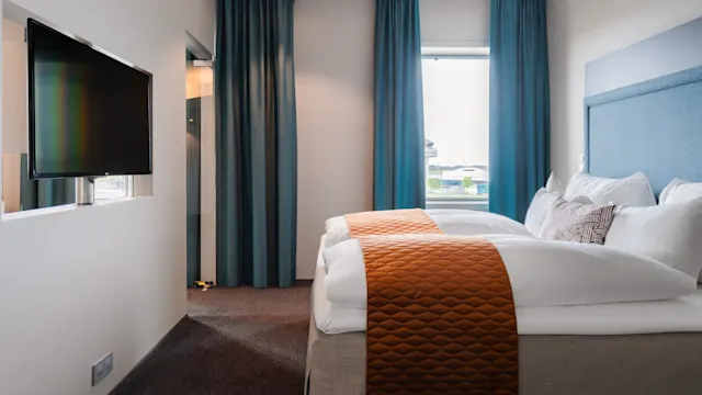 A neatly made bed with orange throws sits against a wall in a hotel room with blue curtains. A flat-screen TV is mounted on the left wall.