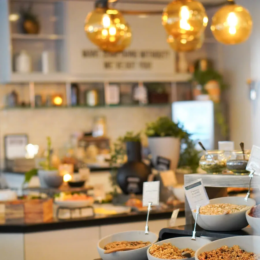 Bowls of cereals sit on a counter, each labeled with a card. In the background, shelves, plants, and warm pendant lights create a cozy, inviting atmosphere. 