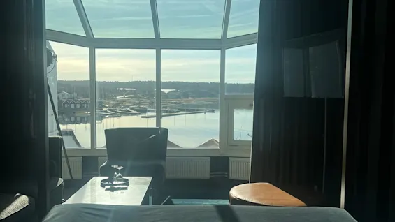 Bed facing large windows overlooking a serene waterfront and buildings, with sun streaming in. A chair and table are positioned in front, with curtains framing the view.