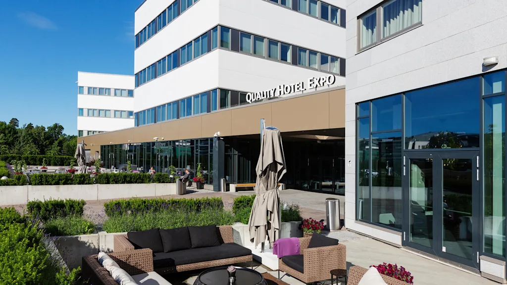 Quality Hotel Expo-bygning med moderne eksteriør, med glass og betong. Utendørs sitteområde med rottingmøbler og parasoller. Omgivende grøntområder og klar blå himmel.