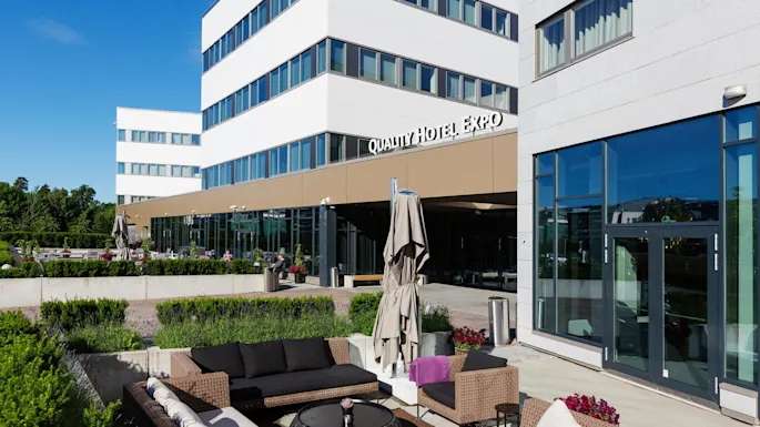 Quality Hotel Expo building with modern exterior, featuring glass and concrete. Outdoor seating area with wicker furniture and umbrellas. Surrounding greenery and clear blue sky. 
