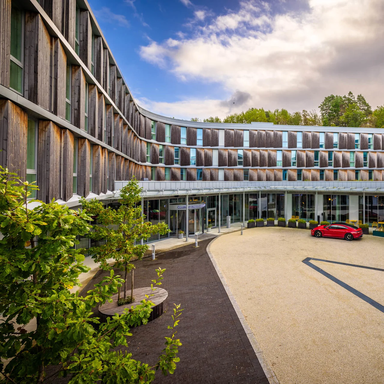 Modern hotel facade with curved wooden panels surrounds a courtyard, featuring a parked red car. Trees and plants add greenery. Signs read "CLARION" and "RESTAURANT."