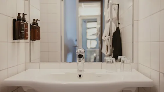 Puhdas valkoinen kaakeloitu kylpyhuone, jossa pesuallas, peili ja SOAP sekä BODY pesuaineannostelijat. Kaksi lasia altaalla.