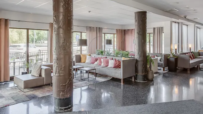 Sitteområde har grå sofaer med fargerike puter rundt trebord, omgitt av store dekorerte søyler og høye vinduer med gardiner. Grønne planter tilfører friskhet til det moderne rommet.