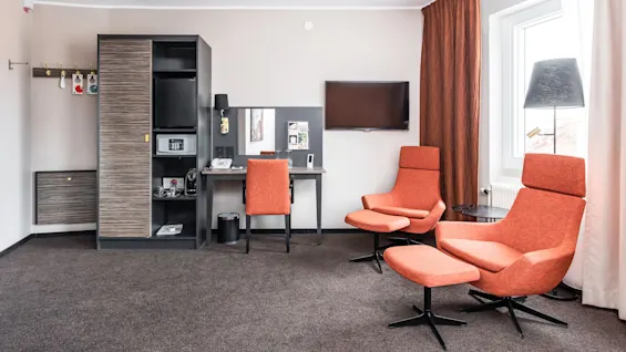 Ett modernt rum med två orange fåtöljer, ett skrivbord, tv och en hyllsektion, antyder ett hotell eller vardagsrum.