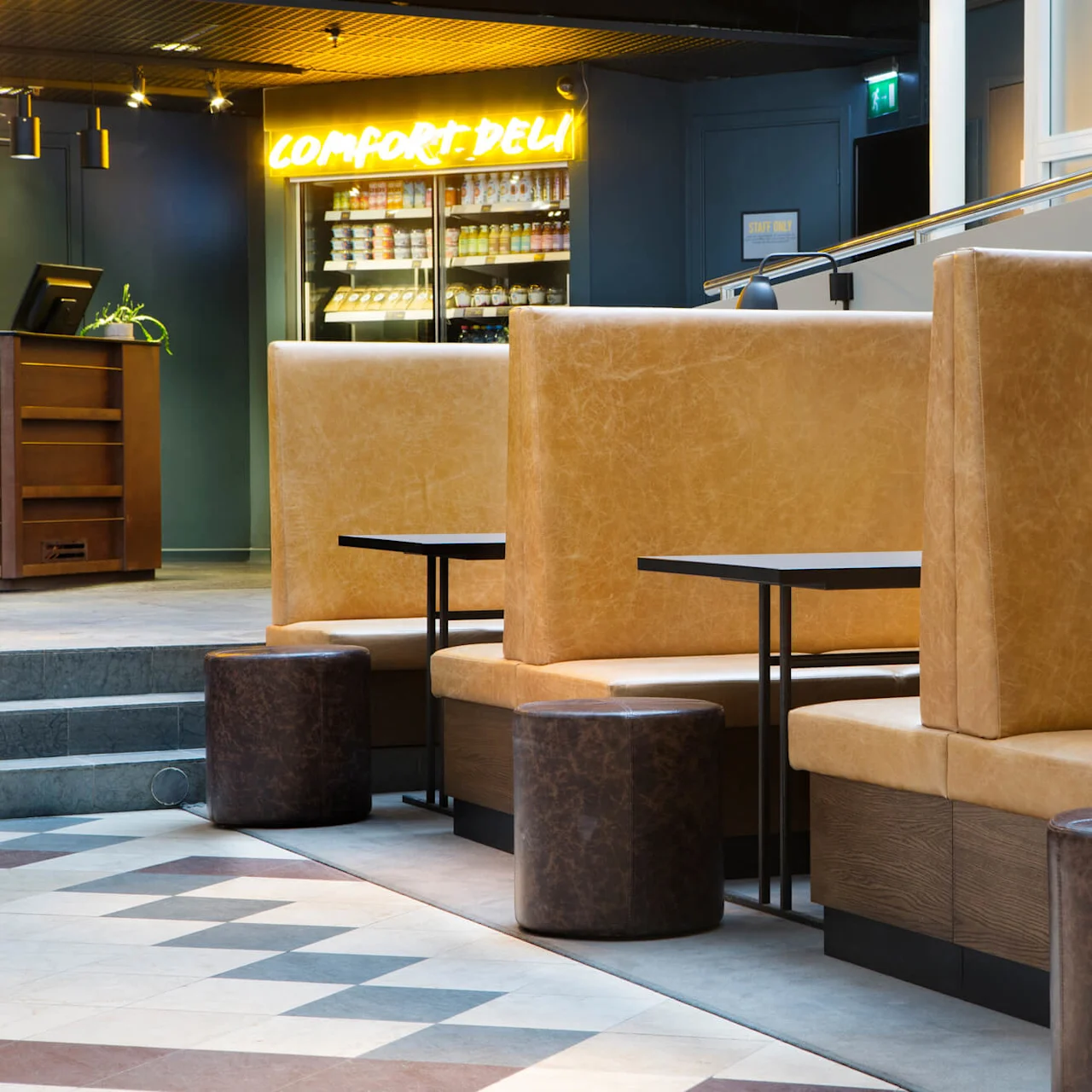Højryggede sofaer og runde skamler i et moderne caféområde; et neonskilt viser "COMFORT DELI".