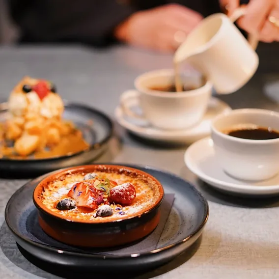 En dessert toppad med bär står på ett bord bredvid två koppar kaffe. En hand häller grädde i en kopp, omgiven av en växt.