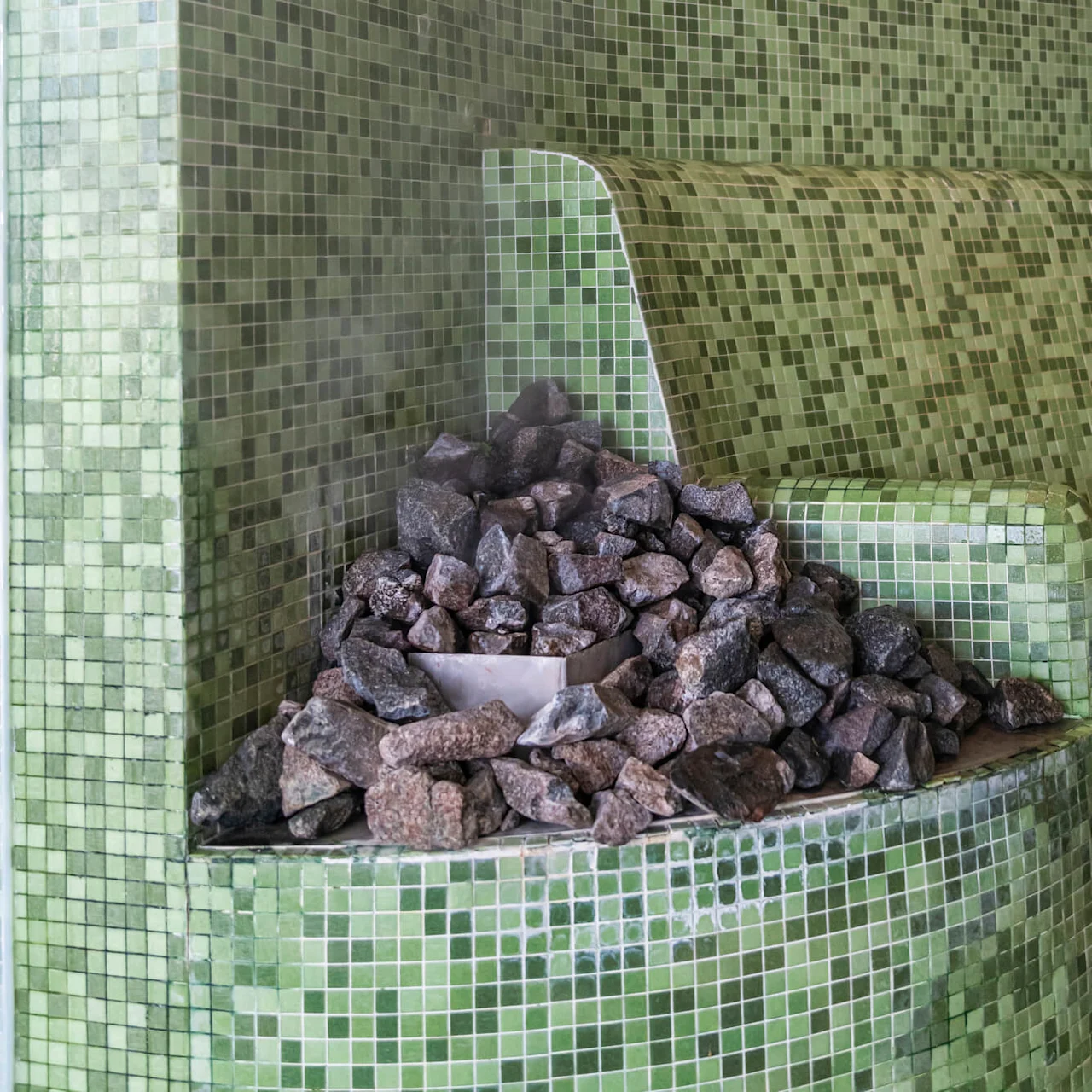 En bunke med dampende varme steiner hviler på en grønn mosaikkflisbenk, og skaper en varm, tåkete atmosfære som er typisk for en badstuomgivelse.