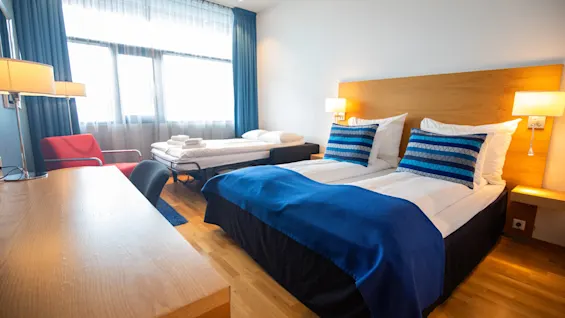 Et hotellrom med to pent oppredde senger med blå detaljer, et skrivebord i tre og en rød stol. Naturlig lys filtrerer gjennom store vinduer med blå gardiner.