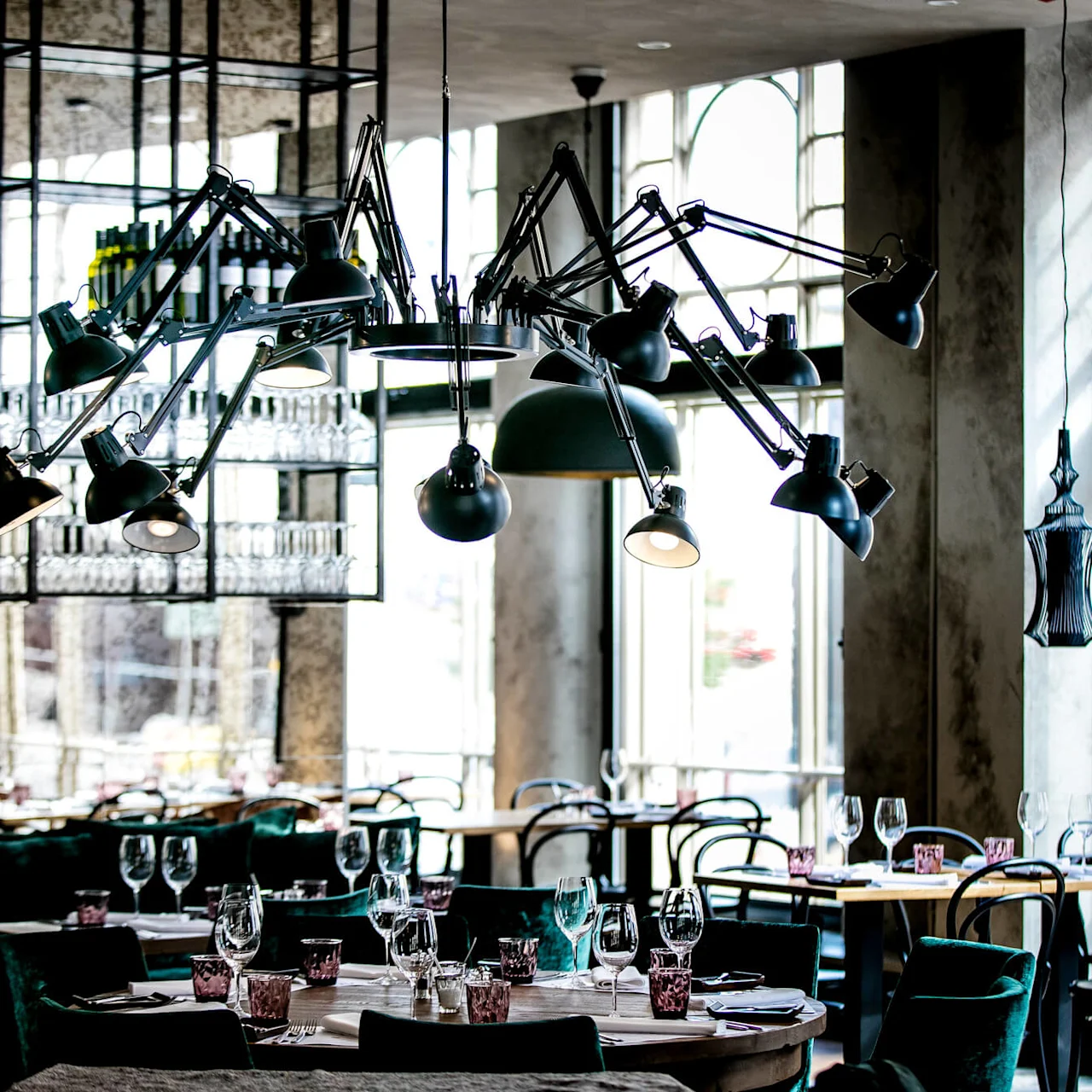 Stor moderne lysekrone med flere justerbare armer henger over elegant oppsatte restaurantbord med vinglass og rosa glass. Solfylt rom med store vinduer og minimalistisk innredning.
