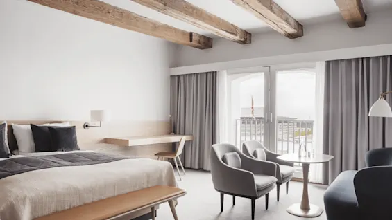 A neatly arranged hotel room features a stylish bed, small round table, two chairs, and a desk. Large windows allow natural light, with visible wooden beams above.