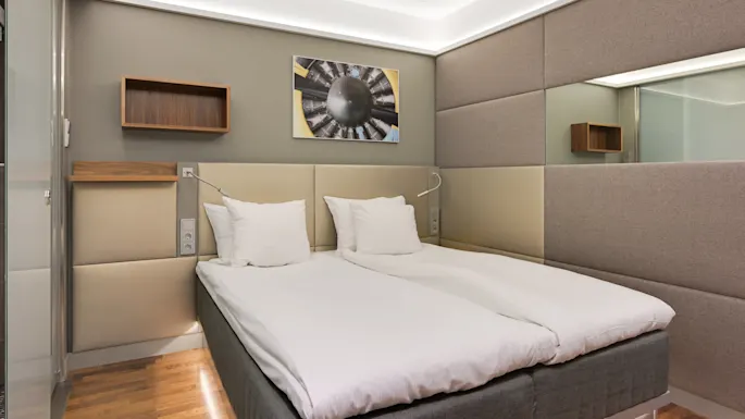En seng med hvitt sengetøy og puter står mot polstrede vegger i et moderne rom med tregulv. Et futuristisk kunstverk henger over sengen.