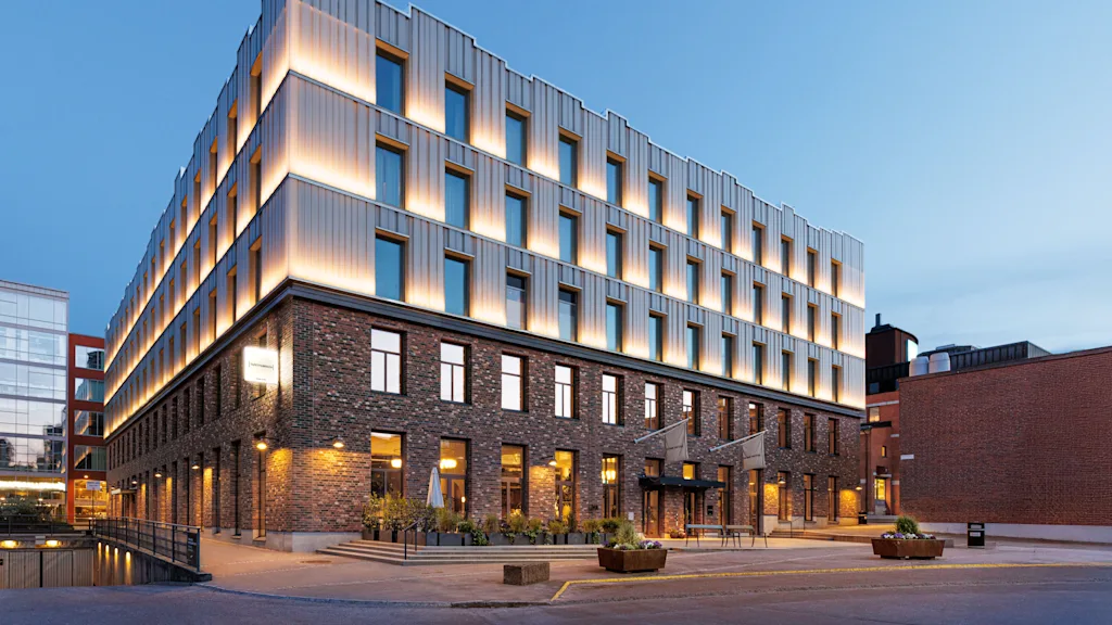 A modern, multi-story building with illuminated upper sections and brick lower levels stands in an urban setting at dusk, surrounded by a paved area and neighboring structures.