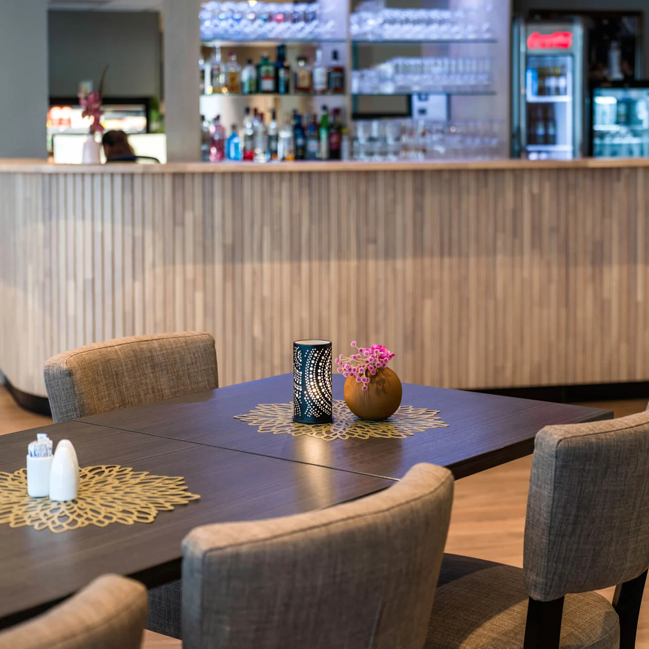 A table with decorative mats holds a vase of pink flowers and a candle holder, surrounded by upholstered chairs. In the background, a wooden bar displays various bottles.