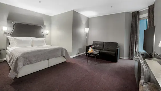 En pænt redt seng med væglamper står i et blødt oplyst hotelværelse, sammen med en sort sofa og et bord, og et stort TV til højre.