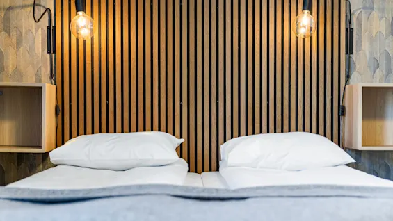 Dobbeltseng med en moderne spilevegg som sengegavl og tente lamper i et hotellrom.