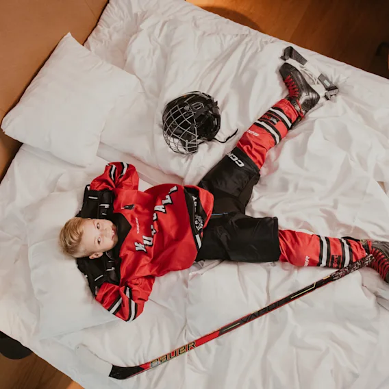 Barn i rød hockeyuniform og skøyter ligger på en seng med hjelm og kølle, tar en pause.