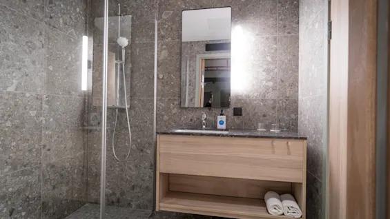 Shower enclosed with glass; adjacent to sink with a mirror and a wooden cabinet. Bathroom has stone-tiled walls and floor, with neatly rolled towels on the shelf.