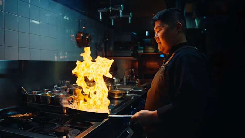A chef holds a flaming pan, creating a dramatic fire display, in a dimly lit professional kitchen with tiled walls and various cooking pots around.