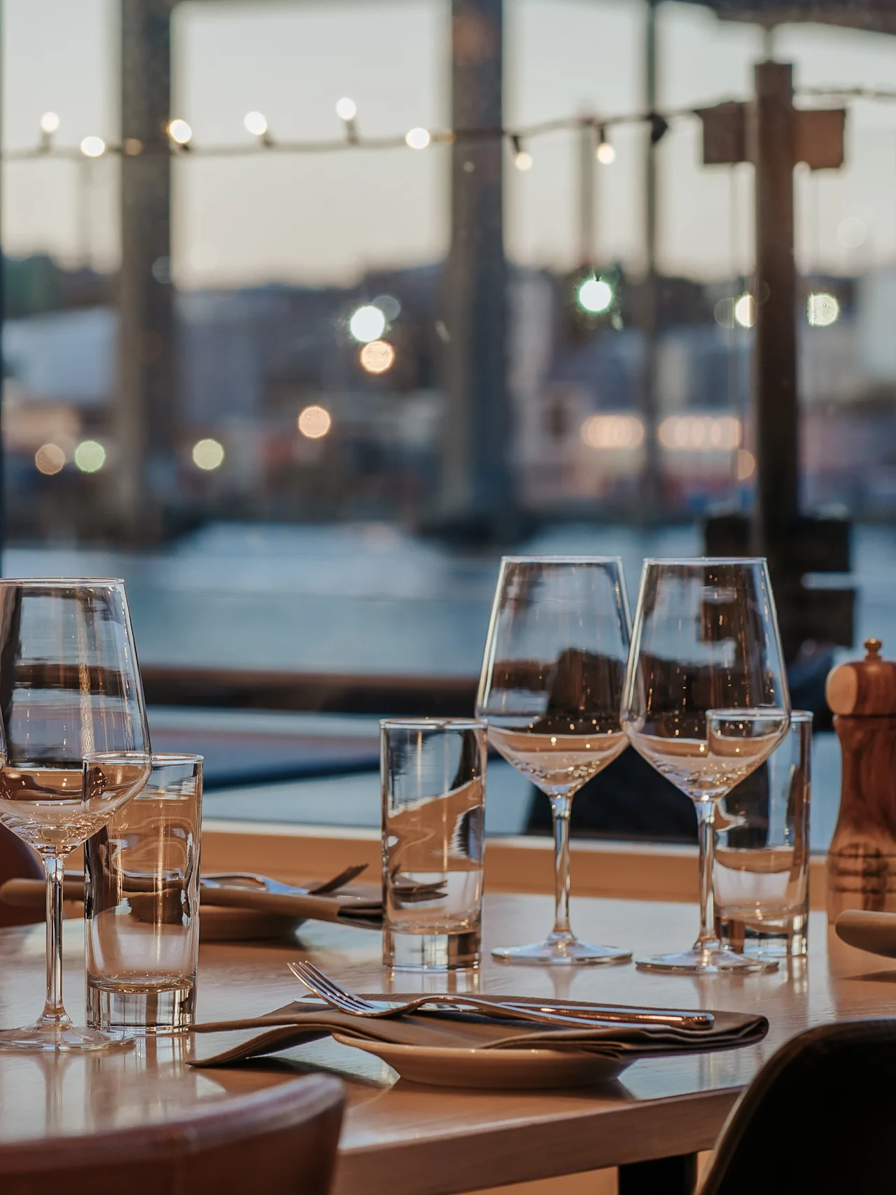 A dining table set with glassware and cutlery sits by a window overlooking a waterfront, with a bridge visible in the background. The atmosphere is softly lit and elegant.