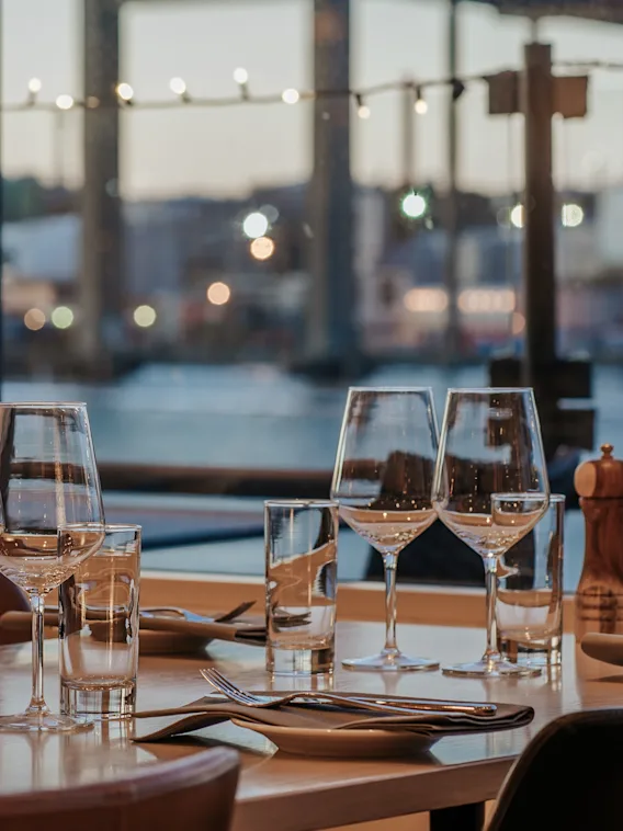 Ett dukat matbord med glas och bestick står vid ett fönster med utsikt över vattnet, med en bro i bakgrunden. Atmosfären är mjukt upplyst och elegant.