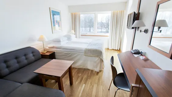 A bed neatly made with white linens sits beside a window in a bright, modern hotel room, featuring a grey sofa, wooden desk, chair, and decorative lamps.