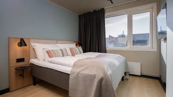 Ett modernt och inbjudande hotellrum med en bekväm säng, randiga kuddar och ett stort fönster som släpper in naturligt ljus.