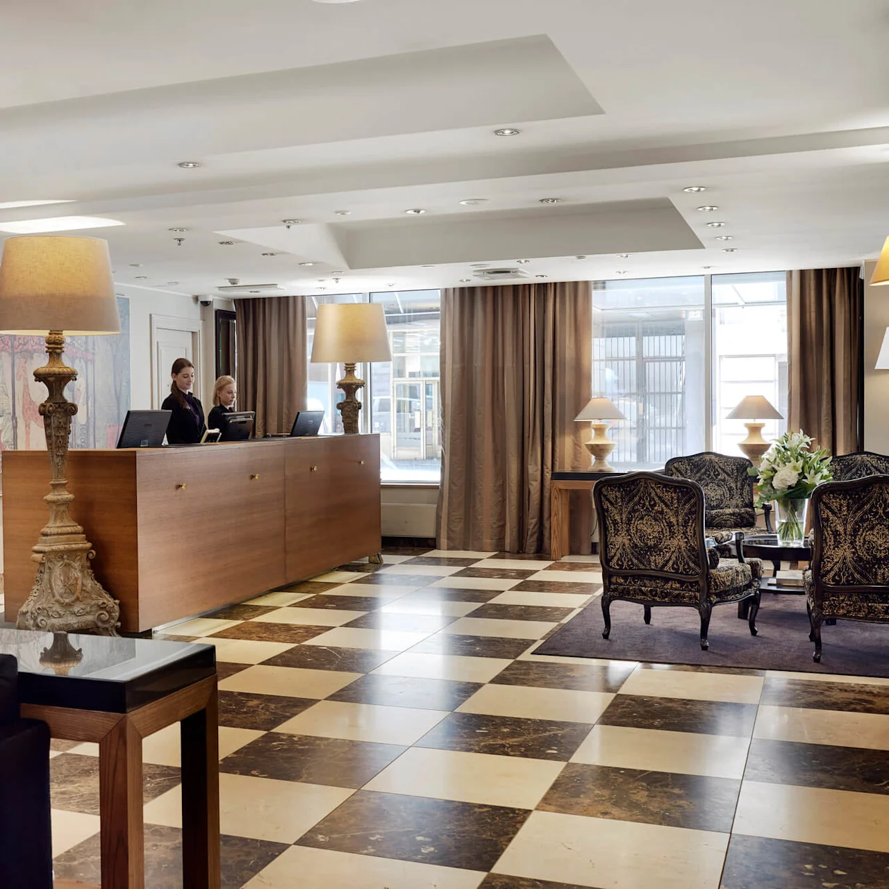 Reception desk with two staff members standing behind, in a hotel lobby featuring decorative lamps, a large floral arrangement, patterned chairs, and checkered flooring.