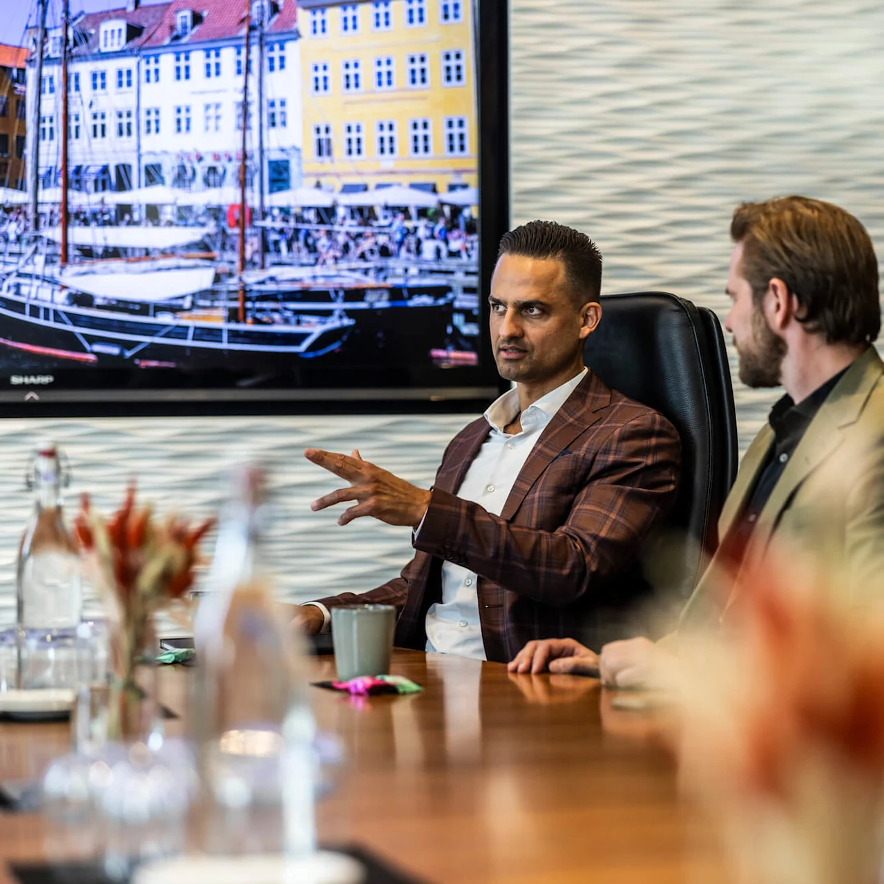 En man i kostym gestikulerar medan han talar under ett möte, omgiven av kollegor. En stor skärm visar en färgglad bild av byggnader vid vattnet och en båt.
