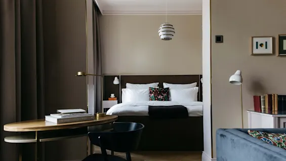 En seng med hvitt sengetøy og fargerike puter er sentrert under en moderne pendellampe. I forgrunnen står et trebord med bøker, inkludert "THE DESIGN HOTELS™ BOOK".
