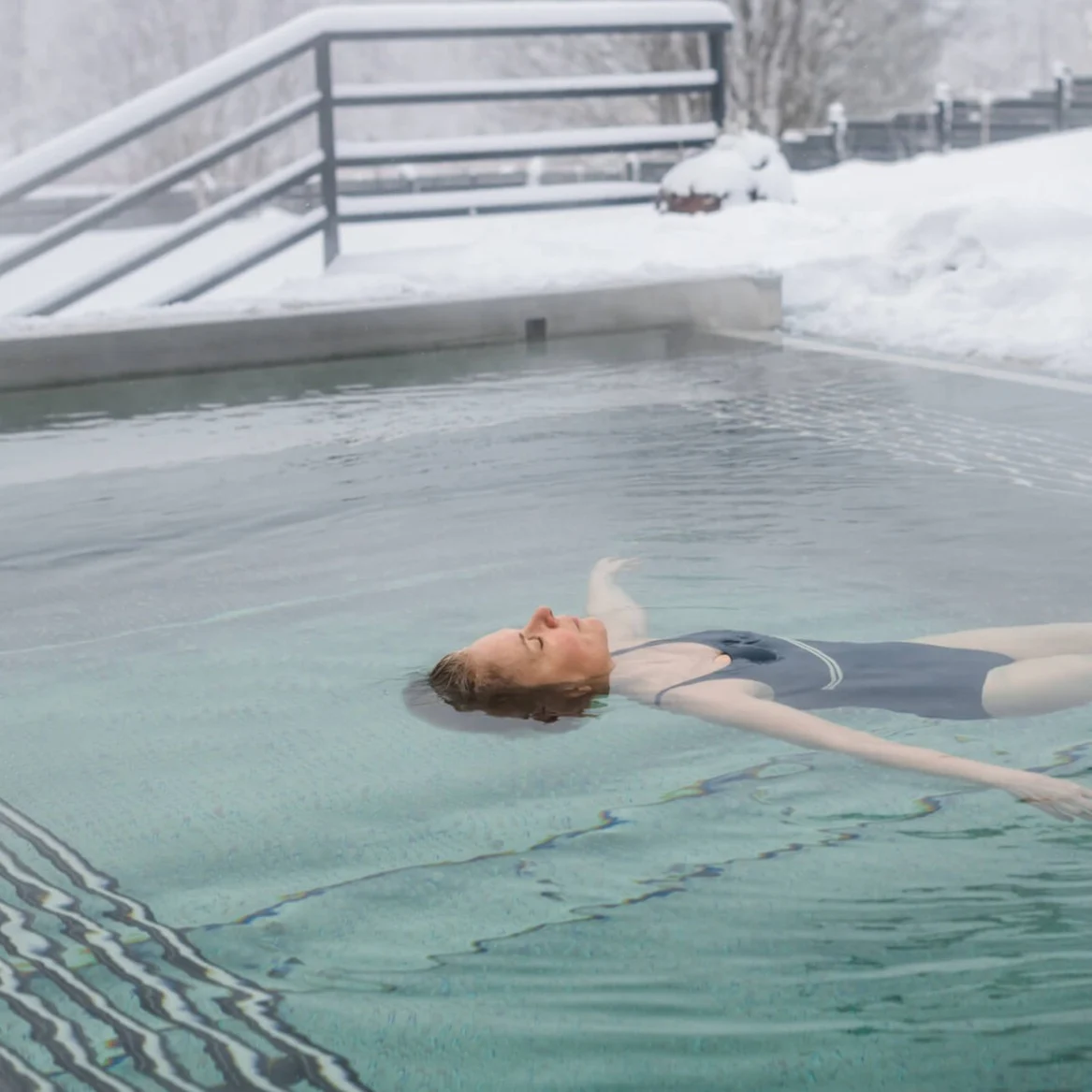 A person floats on their back in an outdoor pool while snow surrounds the area, with bare trees and a railing in the background.