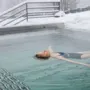 A person floats on their back in an outdoor pool while snow surrounds the area, with bare trees and a railing in the background.