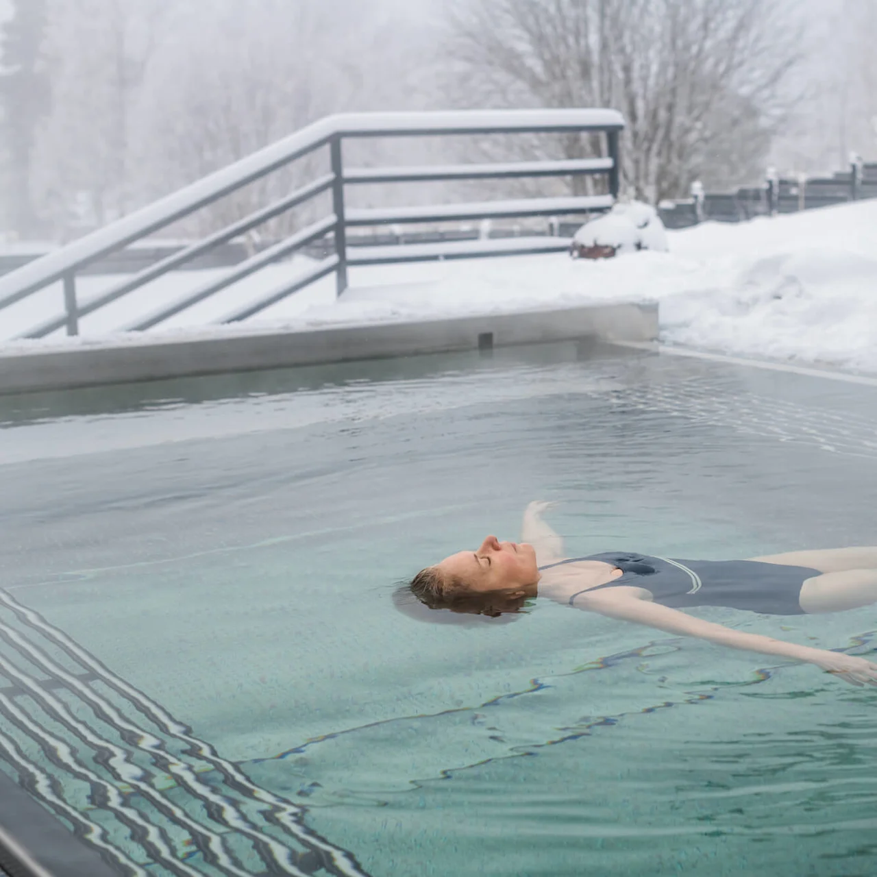 En person flyter på rygg i en utomhuspool medan snö omger området, med kala träd och ett räcke i bakgrunden.