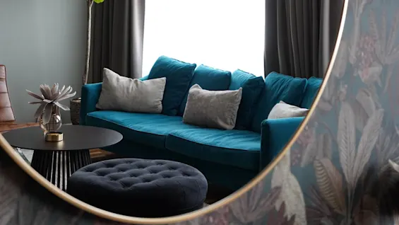 A teal sofa adorned with gray cushions faces a round ottoman. A plant stands beside, while dark curtains frame the bright window, reflecting floral wallpaper in a nearby mirror.
