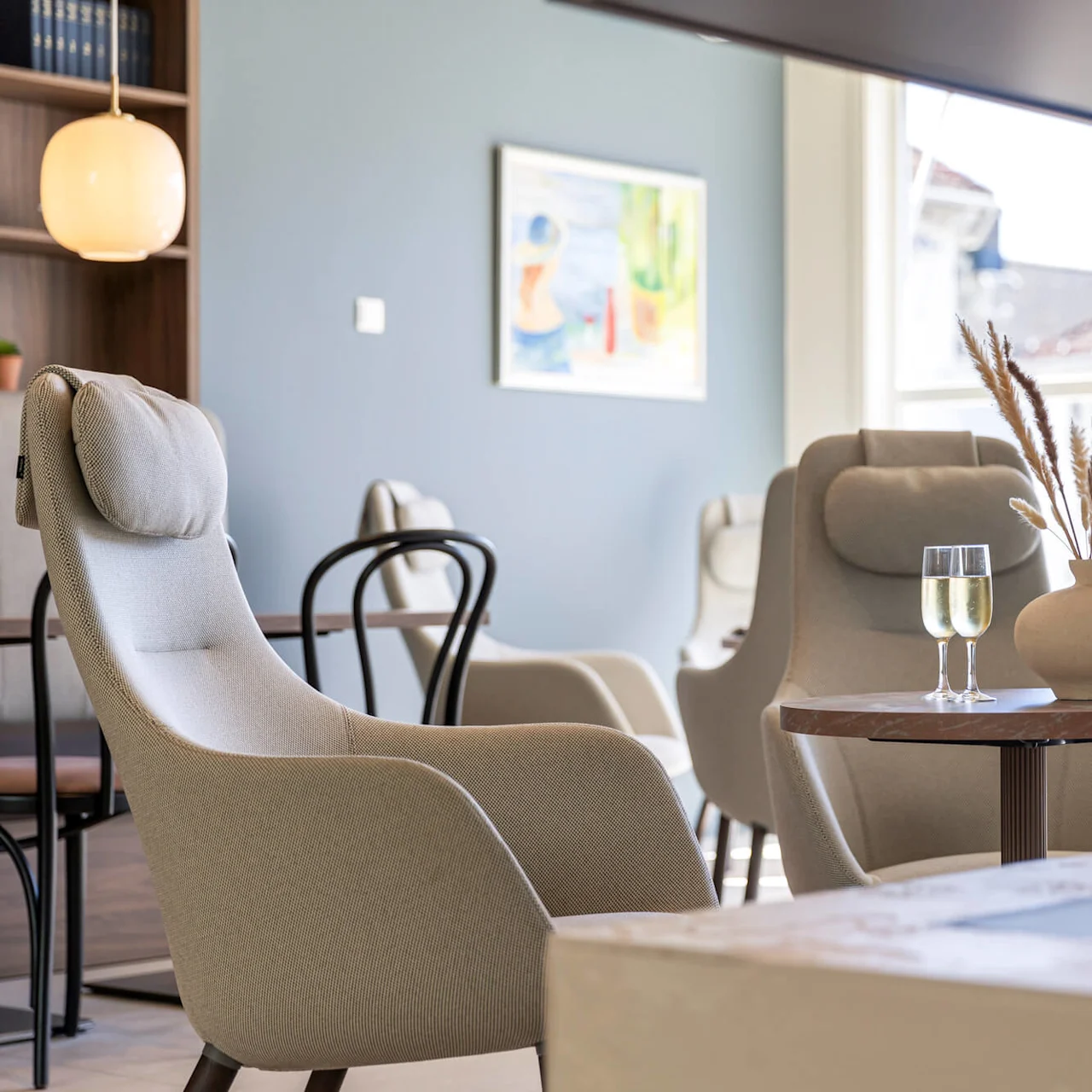 Modern furniture in a well-lit interior space with large windows and art on the wall; water glass on a table beside a comfy chair.