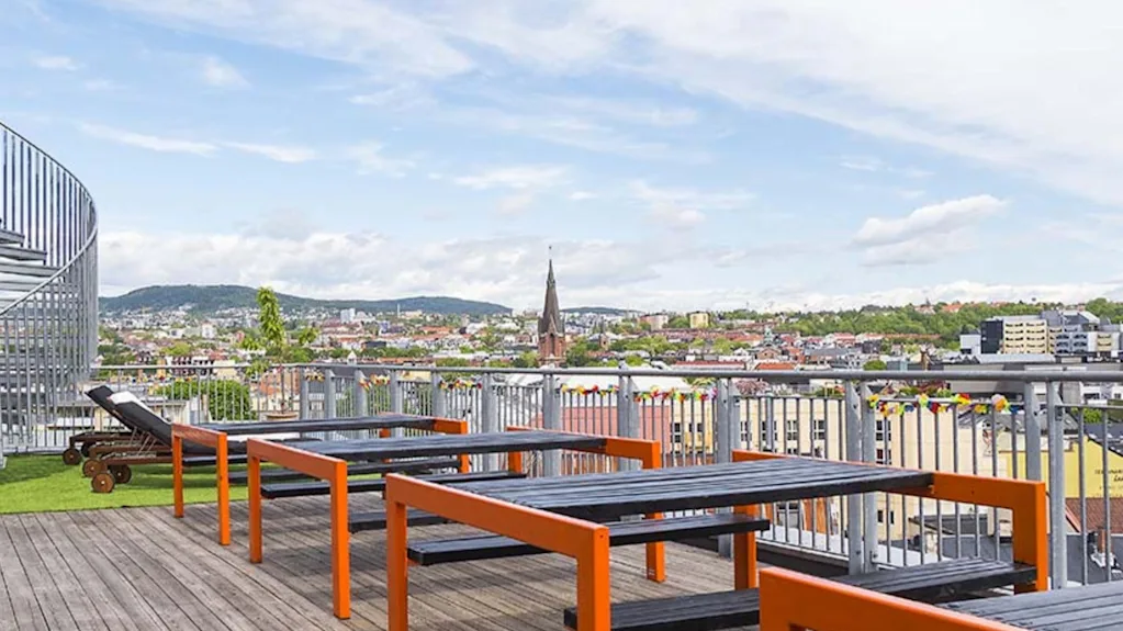 Træborde med orange rammer står på en tagterrasse og kigger ud over en bysilhuet med et kirketårn og fjerne bakker under en delvist overskyet himmel.