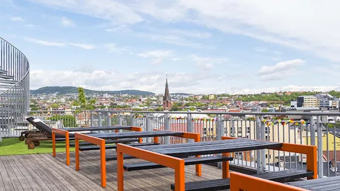 Wooden tables with orange frames stand on a rooftop terrace, overlooking a cityscape with a church spire and distant hills under a partly cloudy sky.