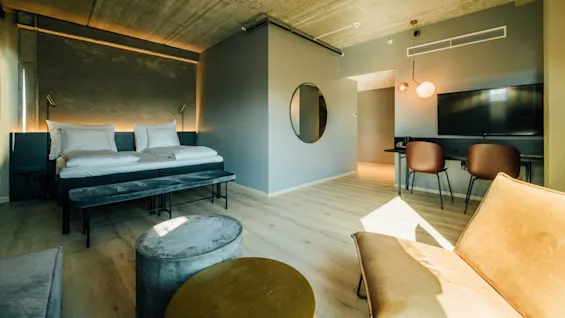 Et hotelværelse med to senge, et arbejdsområde med stole og en TV. Moderne møbler inkluderer en bænk og pouf. Blød belysning kommer bag sengene.
