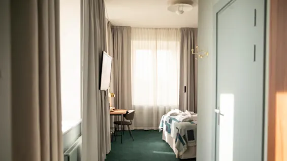 Ett snyggt ordnat rum med en säng täckt av en mönstrad filt, ett skrivbord med en stol och mjukt naturligt ljus från ett gardinfönster. Atmosfären är lugn och mysig.