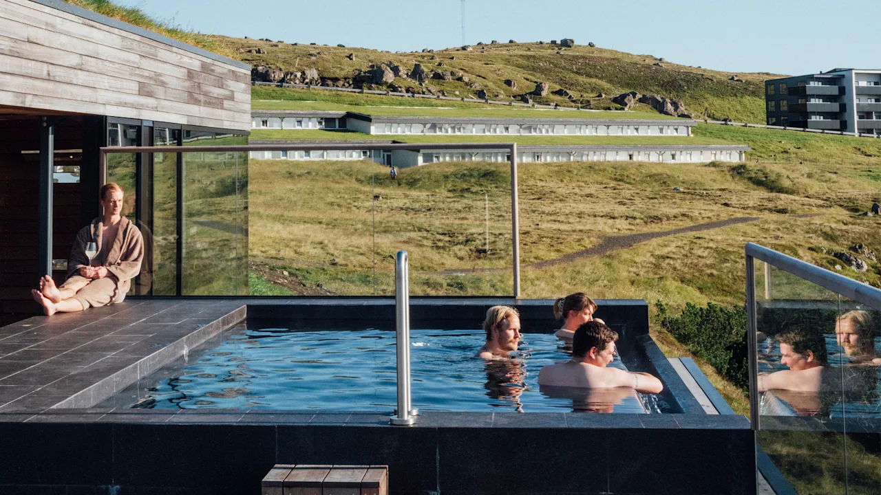 People enjoying an outdoor pool with a scenic view of a grassy hillside and modern buildings.