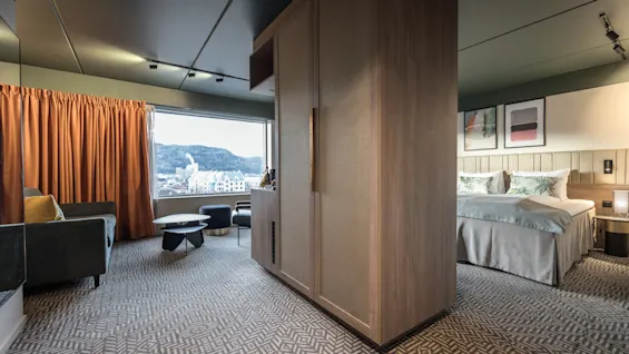 Hotellihuone, jossa parivuode, koristeelliset tyynyt ja abstraktia taidetta seinällä. Oleskelualue, jossa sohva ja pöytä, ison ikkunan vieressä vuoristonäkymällä.