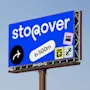 Mainostaulu näyttää "stopover" sinisellä taustalla. Kuvakkeet sängystä, pesukoneesta ja käsipainosta. Nuoli oikealle tekstillä "In 500m." Kirkasta taivasta vasten.