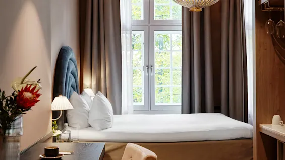 A neatly made bed with white pillows is placed beside a window with grey curtains in a cozy room. A wooden desk holds a vase with flowers and a cup. 
