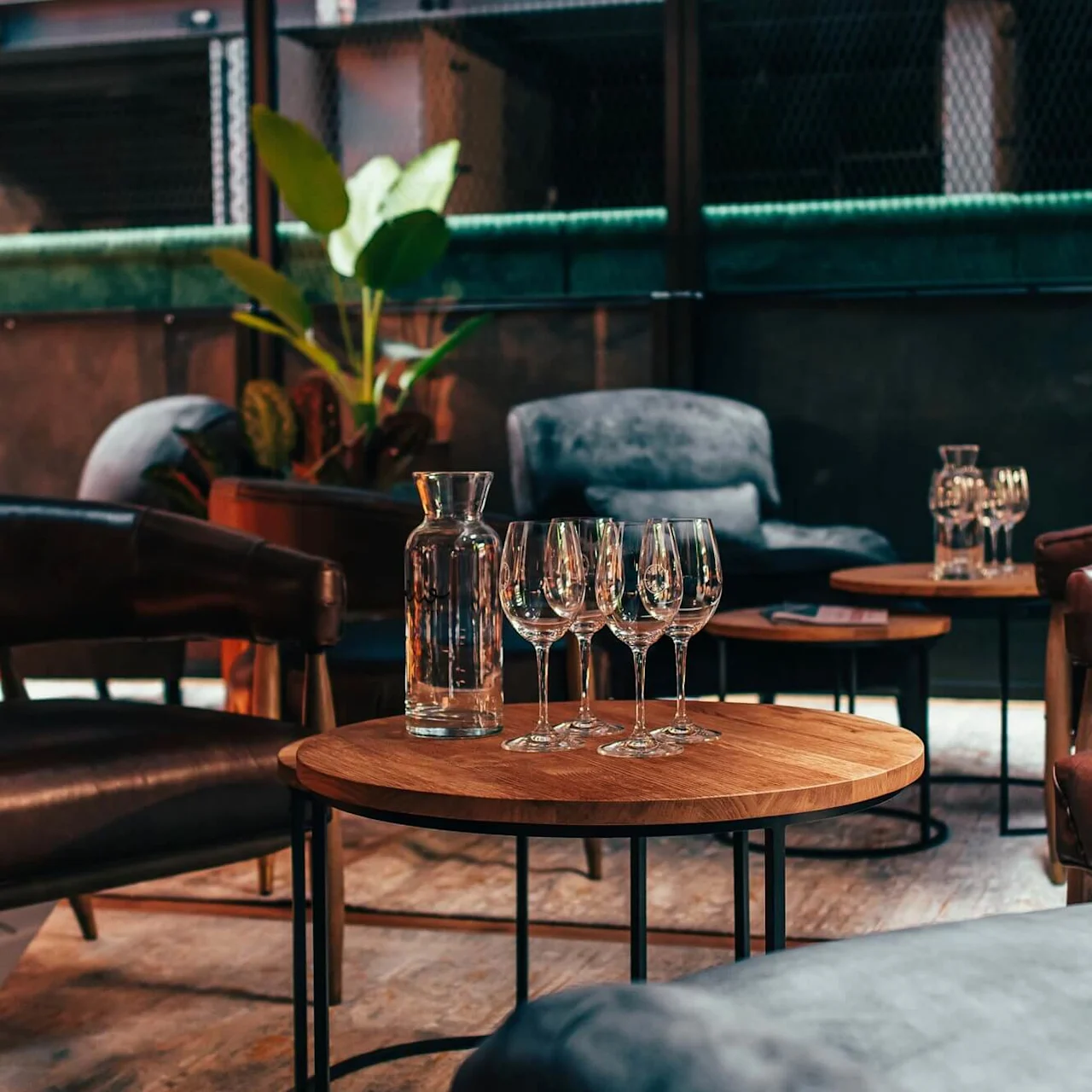 En karaff og vinglass hviler på et trebord omgitt av skinnstoler i en moderne, mykt opplyst lounge. Høye grønne planter og koselige sitteplasser skaper en innbydende atmosfære.