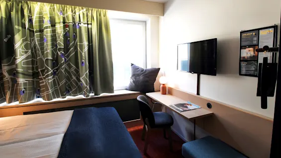 Et hotellrom med en seng, en veggmontert TV, et skrivebord med lampe og magasin, nær et vindu med designgardiner. Tekst: "HELP?", "THIS IS HOW IT WORKS", "WE CARE".