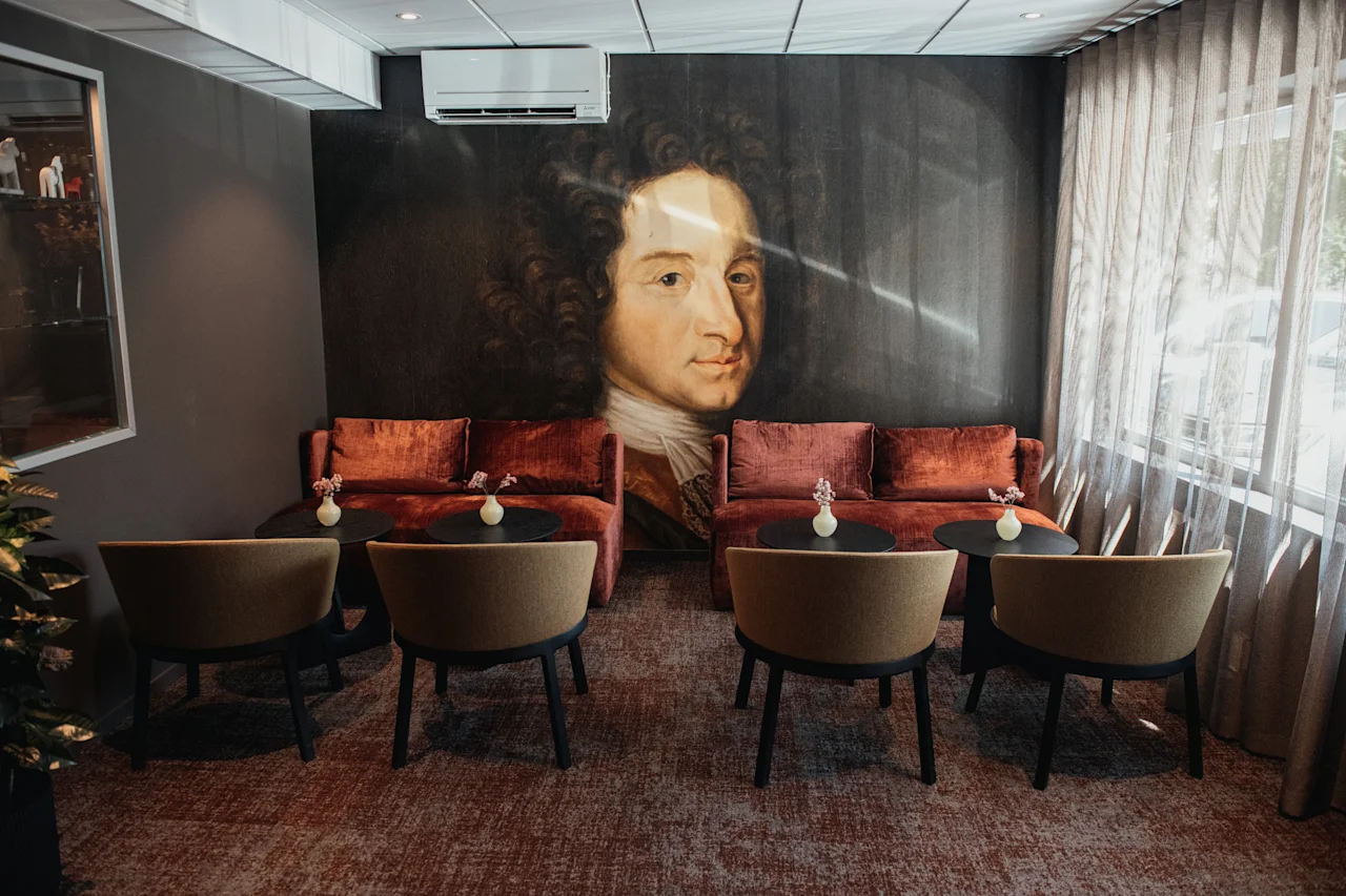 Et koselig salongområde med røde sofaer, stoler og små bord, med et stort portrettmaleri på veggen og naturlig lys fra et vindu.