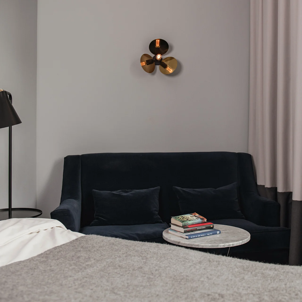 En marinblå soffa står mot en grå vägg under en modern vägglampa. Ett litet runt bord med böcker står i närheten. Gardiner täcker delvis väggen bredvid en säng.