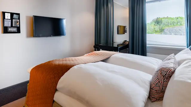 Sänky sijaitsee modernissa hotellihuoneessa, jossa on seinälle kiinnitetty televisio, puhelin pöydällä ja ikkunasta näkyy vehreä ympäristö. Seinillä on kuvia.
