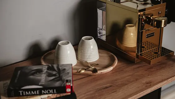 A golden coffee machine by "SJÖSTRAND" sits on a wooden counter next to two upside-down mugs on a tray, accompanied by golden spoons, with books titled "TIMME NOLL."
